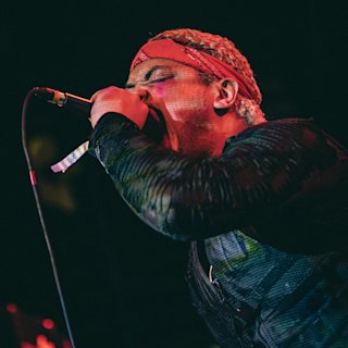 Artist Kid Bookie wearing a red bandana passionately sings into a microphone on stage, illuminated by stage lighting.