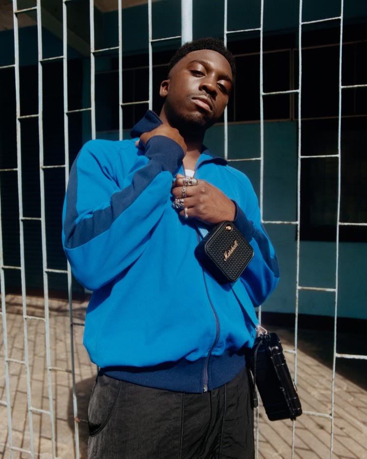 A man wearing a blue zip-up jacket and holding Marshall Willen II Black and Brass portable Bluetooth speaker.