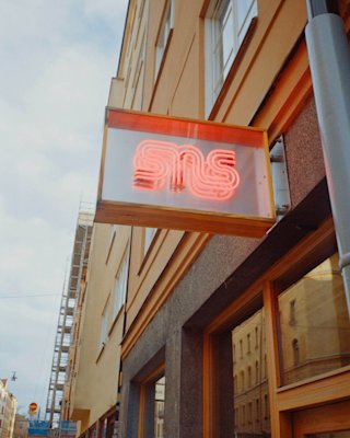 Cartel de neón con las letras "sms" montado en el exterior de un edificio en un día soleado, con un tenue reflejo y un cielo azul despejado de fondo.