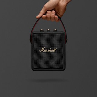 A hand holds Marshall Stockwell III Black and Brass portable speaker.