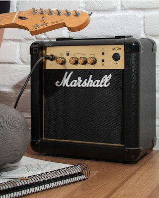Marshall MG10 amplifier, with its connected cable, is set on a wooden floor beside a guitar neck and a notebook.
