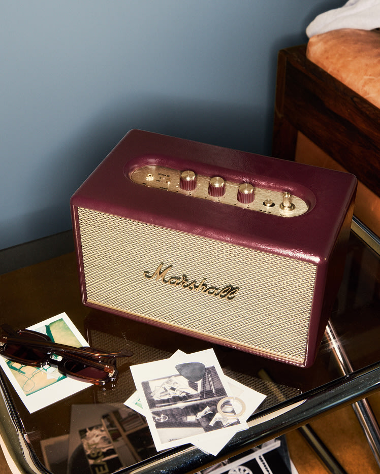 Acton III Burgundy speaker with gold controls sits on a glass table beside sunglasses and black-and-white photos.