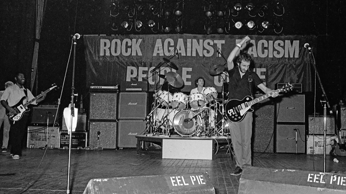 Schwarz-Weiß-Foto eines Gitarristen und eines Schlagzeugers, die bei einem Rock-gegen-Rassismus-Konzert auf der Bühne stehen, mit Transparenten und Bühnenausrüstung.
