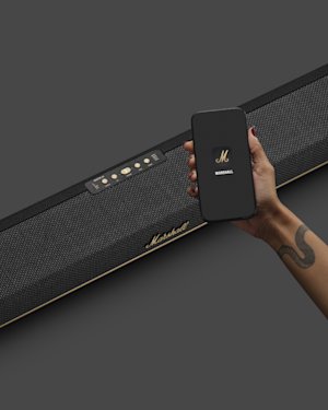 Una mano sostiene un teléfono que muestra la aplicación Marshall frente a una barra de sonido Marshall Heston 60 TV negra.