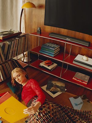 A woman lies on a wooden floor with vinyl records beside her, near the Marshall Heston 60 soundbar, a shelf, and a turntable in a retro-styled room.