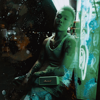 Bohan Phoenix sitting in front of a neon light display with a Marshall Emberton II Cream Portable speaker on his lap.