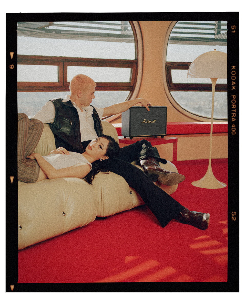 A man and a woman sitting in a sofa beside a Marshall WOBURN III speaker. 