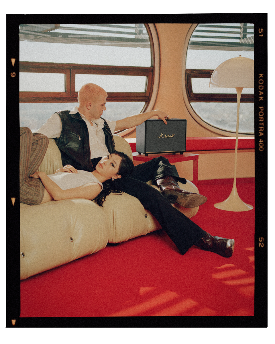 A man and a woman sitting in a sofa beside a Marshall WOBURN III speaker.