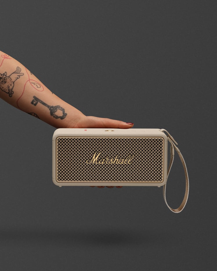 A tattooed hand holds the Marshall Middleton II Cream portable Bluetooth speaker against a plain dark gray background.