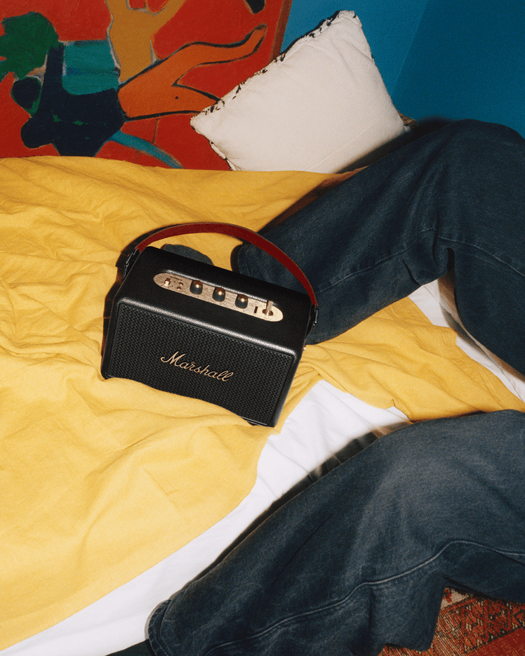 Marshall Kilburn III Black and Brass portable speaker sits on a yellow bedspread next to a person in dark jeans.