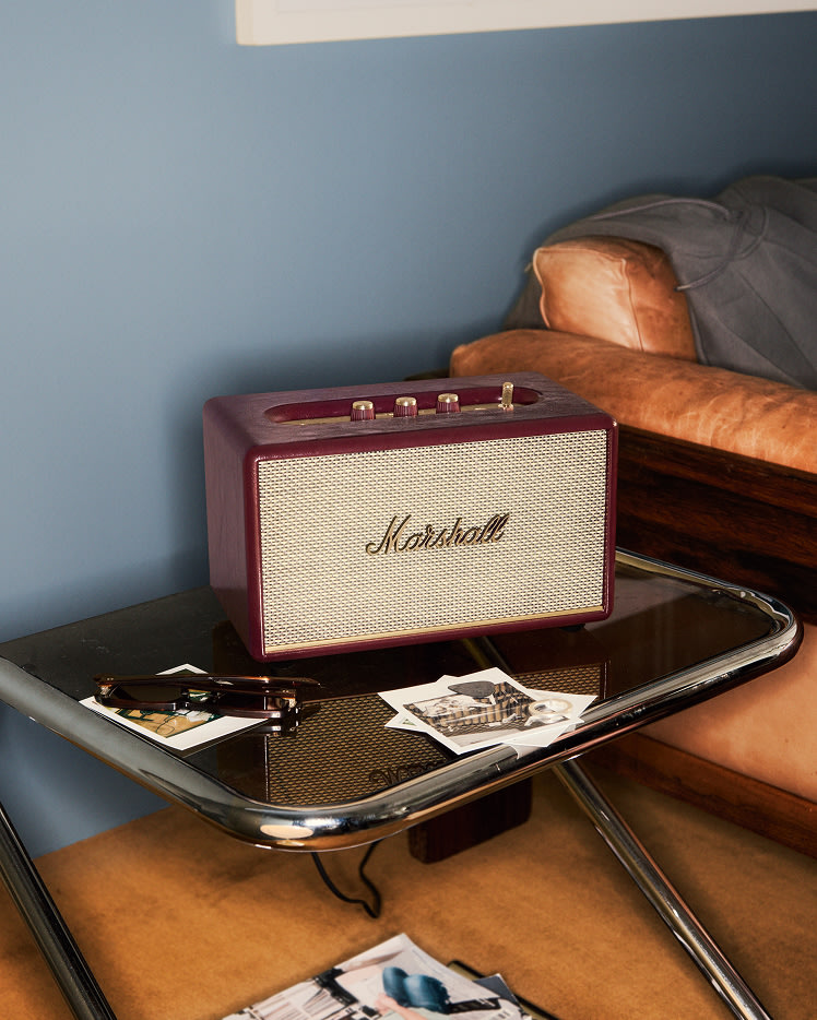 Marshall Acton III Burgundy speaker sits on a glass side table next to a tan leather couch, with scattered photos and sunglasses on the table.