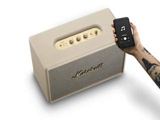 Eine Person hält einen cremefarbenen Marshall WOBURN III-Lautsprecher hoch.