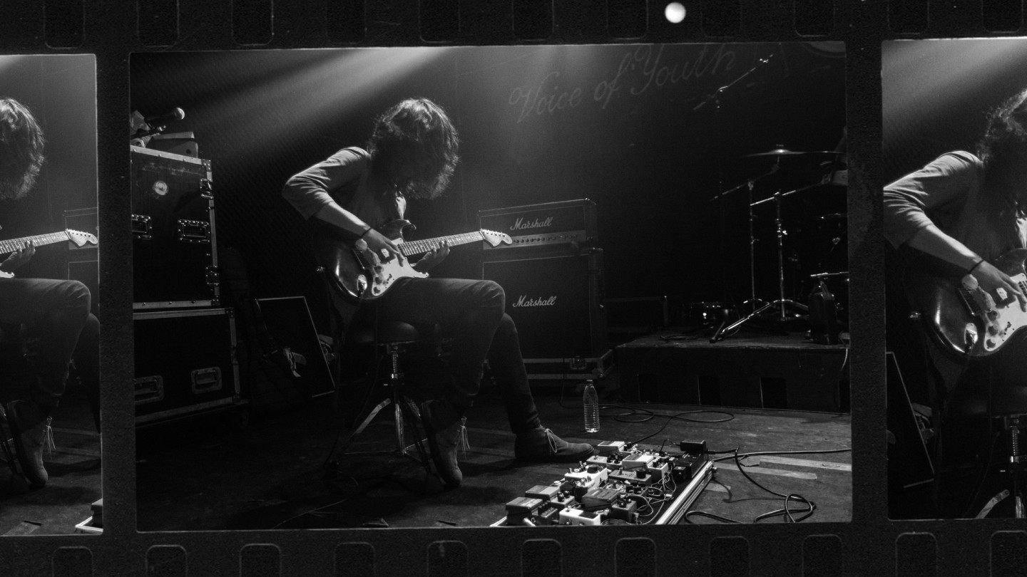 スツールに座り、足元にペダル・ボードを置くギタリストのモノクロ写真。