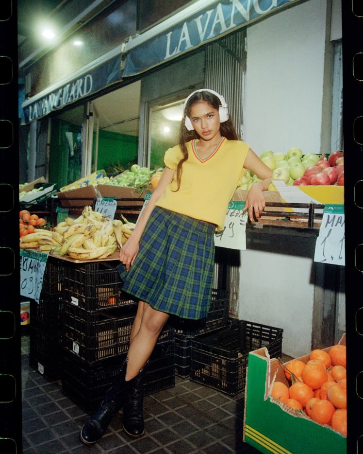 Eine Frau in einem gelben Oberteil und einem karierten Rock steht mit ihren Marshall Major V Cream On-Ear-Kopfhörern in der Nähe eines Obststandes.