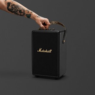 A tattooed arm holds the handle of a Marshall Tufton portable speaker in Black and Brass against a plain dark background.