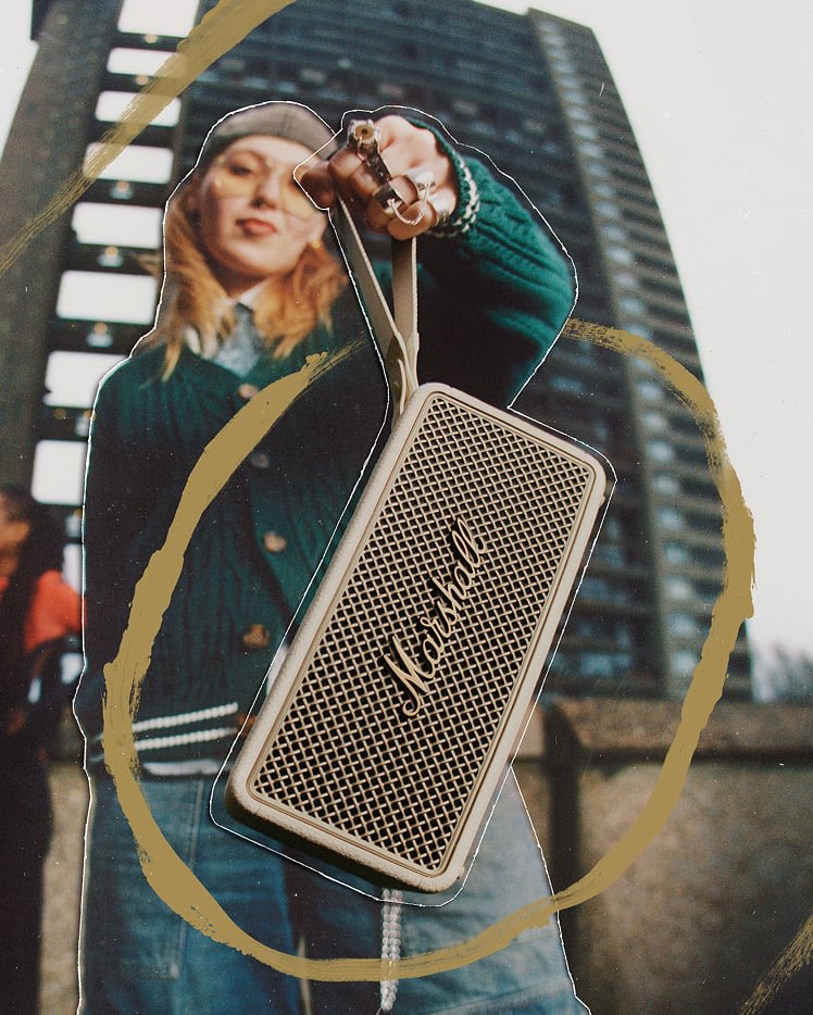 A person holds a Marshall Middleton II Cream portable Bluetooth speaker toward the camera, with a tall building in the background.