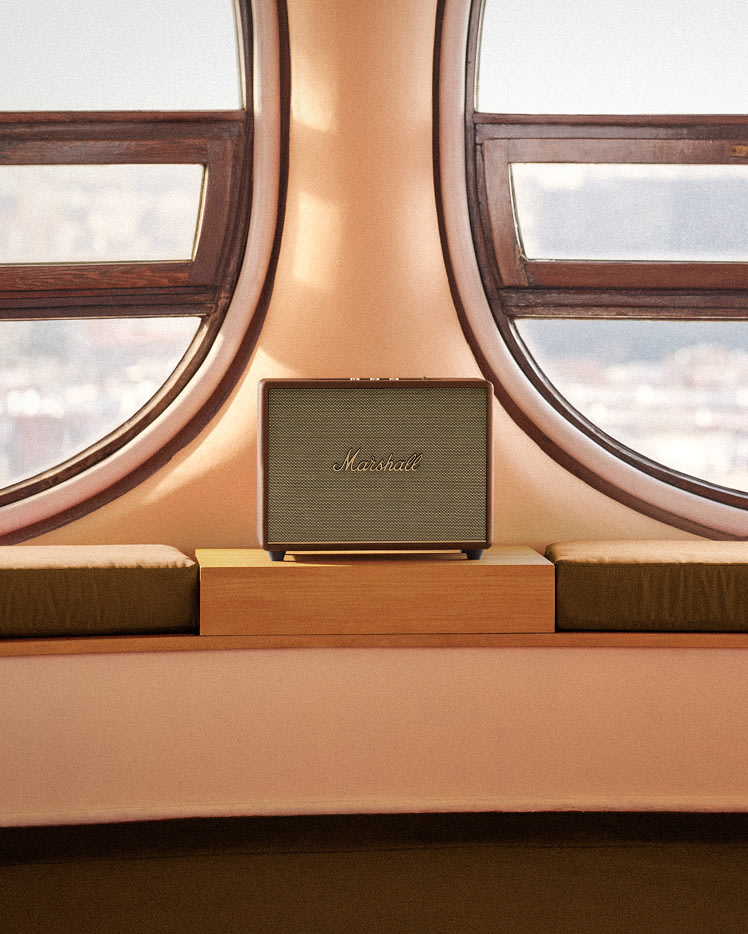 Marshall Woburn III Brown home speaker sits on a wooden shelf between two rounded windows, overlooking a blurred landscape.