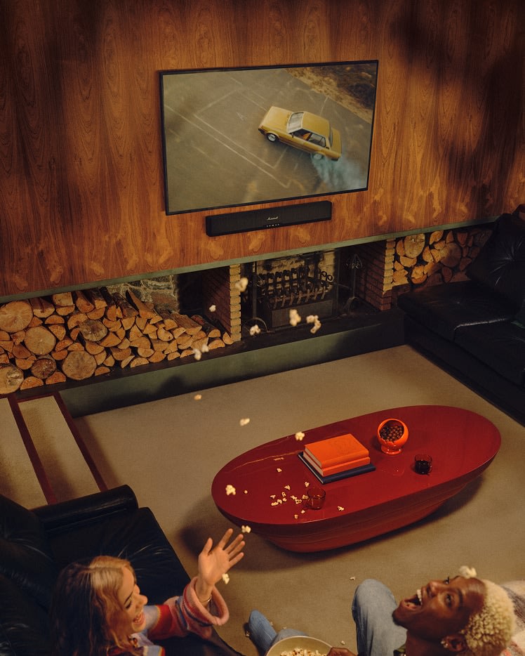 Two people relax on the sofa in a wood-paneled living room, watching a car TV with Marshall Heston 60 Black soundbar.