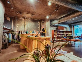 Un hombre detrás de un mostrador de madera curvada en una tienda boutique llena de zapatos y ropa, con decoración de estilo industrial.