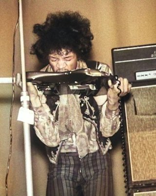 Jimi Hendrix plays a guitar with his teeth onstage, wearing a floral shirt and striped pants, standing next to an amplifier.