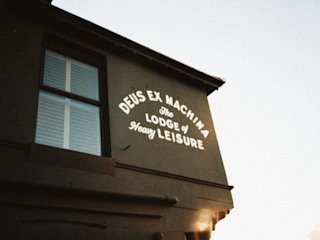 La esquina de un edificio con un cartel que reza "deus ex machina, la logia del ocio pesado" iluminada por la luz del atardecer.