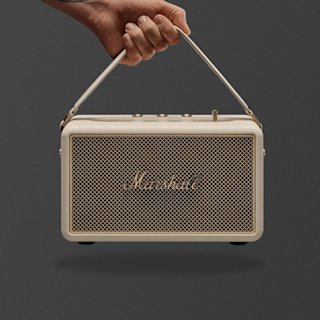 A hand holds a Marshall Kilburn III portable speaker in Cream with a brass fret and control knobs on top against a plain gray background.