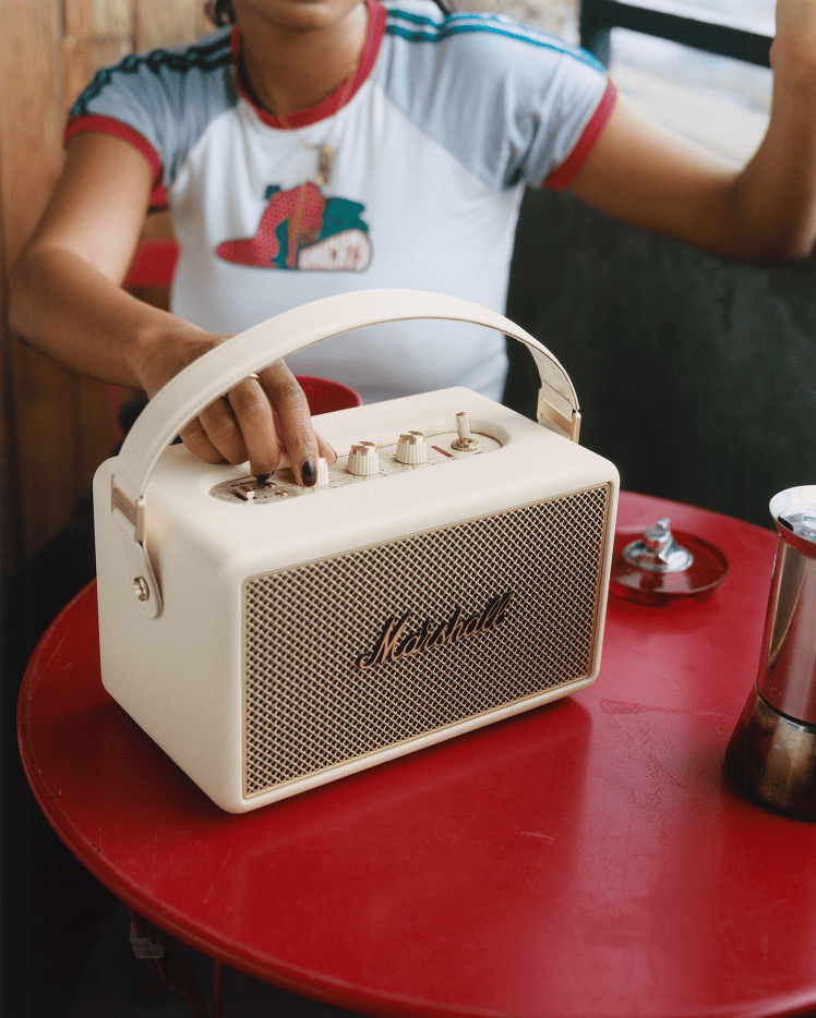A person adjusts the knobs on a Marshall Kilburn III Cream portable speaker, set on a red table.