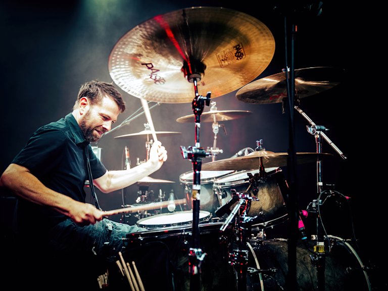 A drummer energetically plays a drum set on a dimly lit stage, surrounded by cymbals.