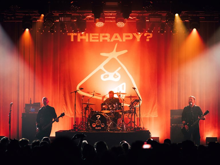 A rock band performs live on stage with "THERAPY?" displayed on a backdrop behind them in large letters. The stage is illuminated with red and orange lights.
