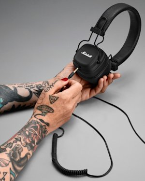 A pair of tattooed hands holding a pair of Major IV headphones, putting a 3,5 mm AUX cable into them.