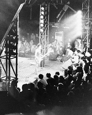 A musician plays guitar on stage under bright lights while an attentive audience watches in a dimly lit venue.