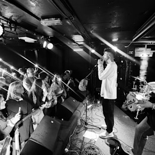 Musician Jarki Monno performs on stage with a microphone, engaging with an audience. The crowd takes photos and videos with their phones while a bassist plays in the background.