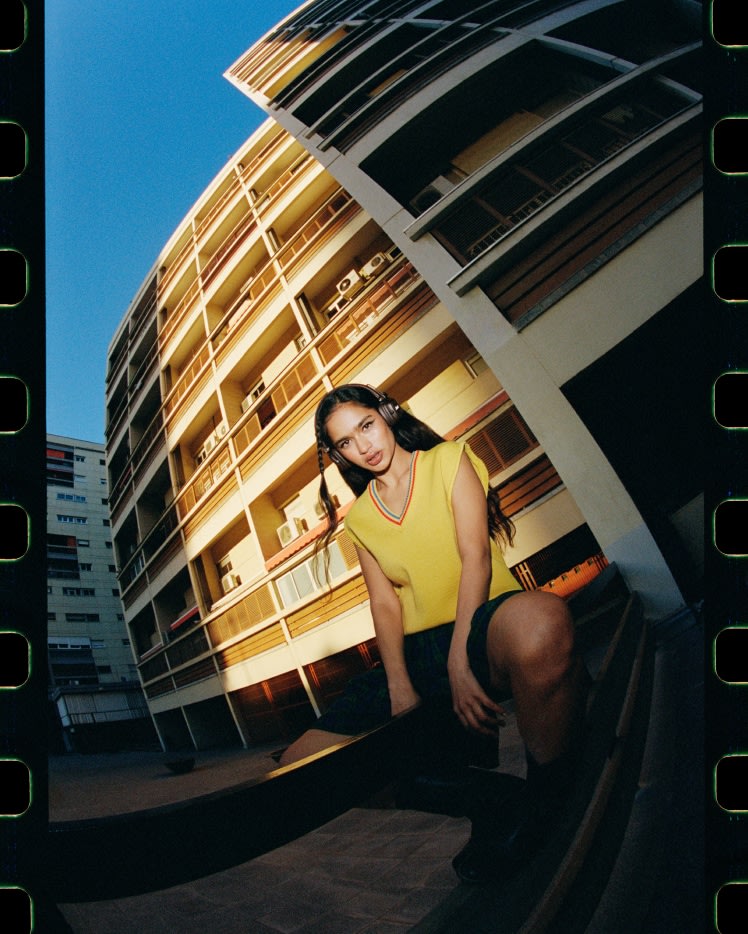 Una mujer con top amarillo y auriculares Marshall Major V Brown on-ear, posa delante de un edificio, con la luz del sol creando sombras.