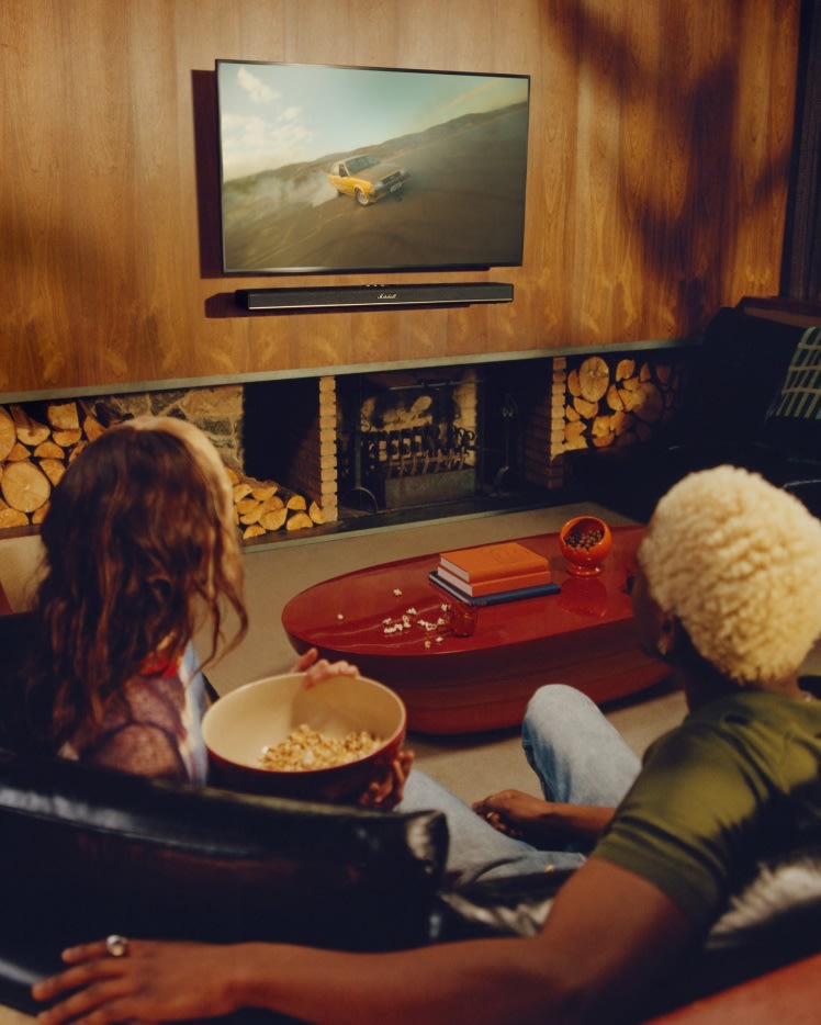 Two people sit on a couch and watching a TV with the Marshall Heston 120 soundbar, in a cozy wood-paneled living room.