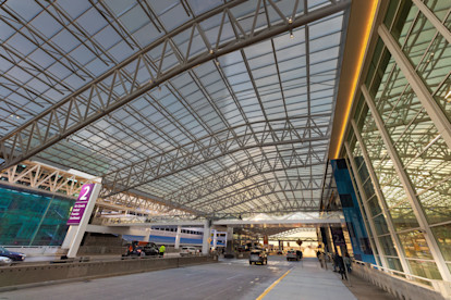 Covered airport terminal drop-off with glass and steel canopy, cars, and passengers at curbside.