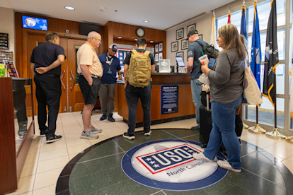 A group of people stands inside a USO Donations center.  The interior features a reception desk with staff assisting visitors, military flags along the right wall, and framed photos and memorabilia on display.