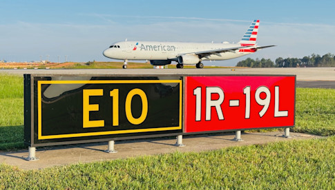 An American Airlines aircraft taxis past a runway identifier sign reading "E10 / 1R-19L" on a grassy airfield under a clear sky.