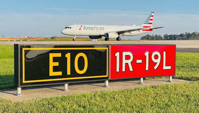 An American Airlines aircraft taxis past a runway identifier sign reading "E10 / 1R-19L" on a grassy airfield under a clear sky.