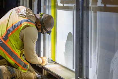 A construction worker in a yellow safety vest, hard hat, and respirator mask crouches to work on the base of a large window or glass panel installation at an indoor job site.
