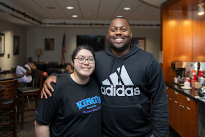Two people smiling and posing together inside a lounge or break area with a U.S. flag in the background.