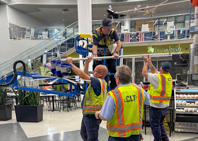 A crew of CLT airport workers in yellow safety vests work together to maneuver a large piece of equipment or signage inside an airport terminal, with a restaurant and escalator visible in the background.