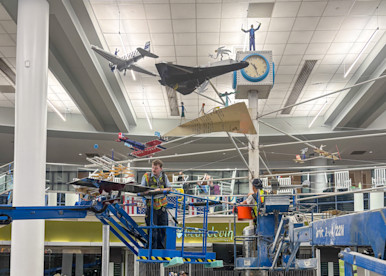 Workers on aerial lift platforms install or restore a large suspended aviation-themed art installation featuring model aircraft, a clock, and a human figure inside an airport terminal.
