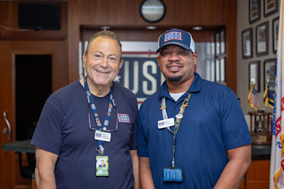 Two USO staff members smile in front of the welcome desk, wearing uniforms