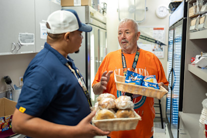 Two USO volunteers talk while holding trays of snacks and pastries in a supply room stocked with food and drinks.