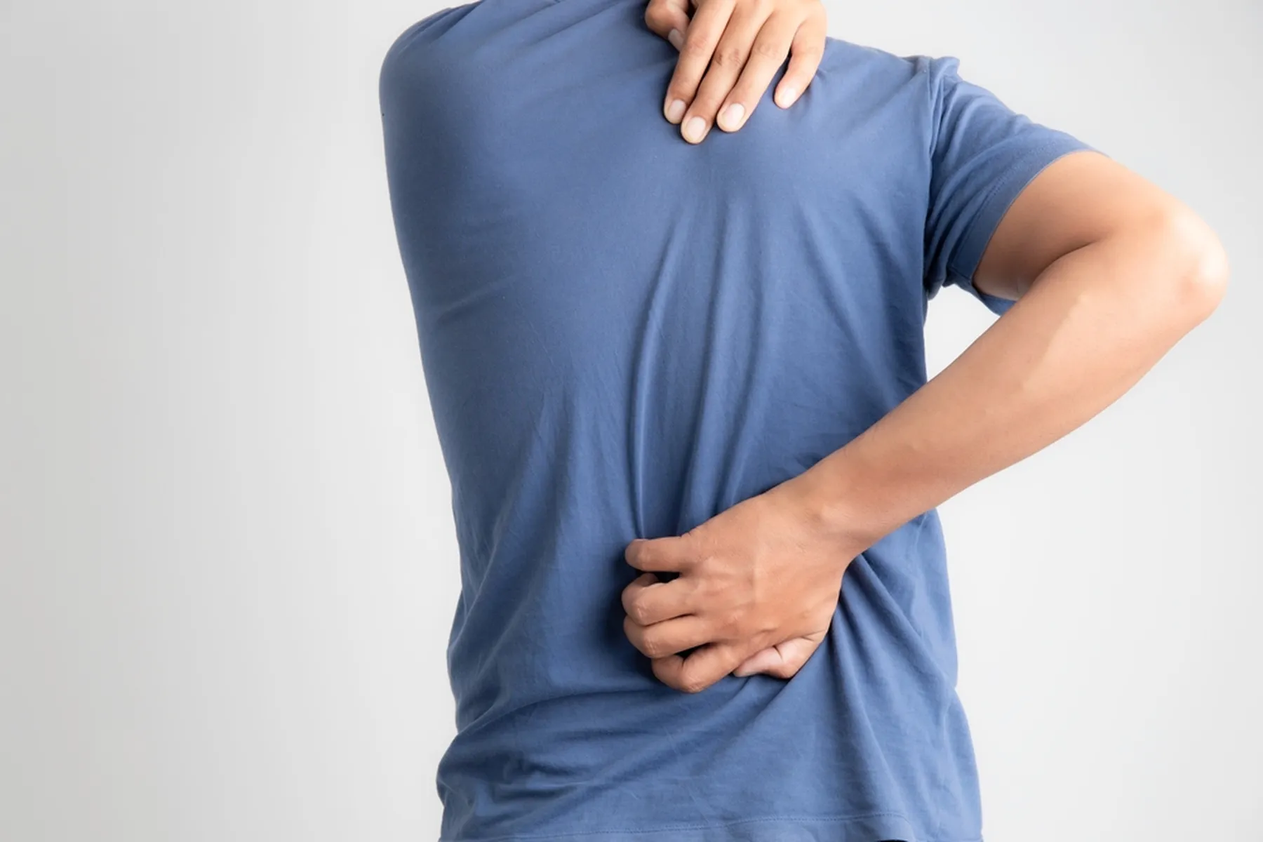 A man's in a blue t-shirt has his back facing the camera as he stretches his hands around and scratches his back. 