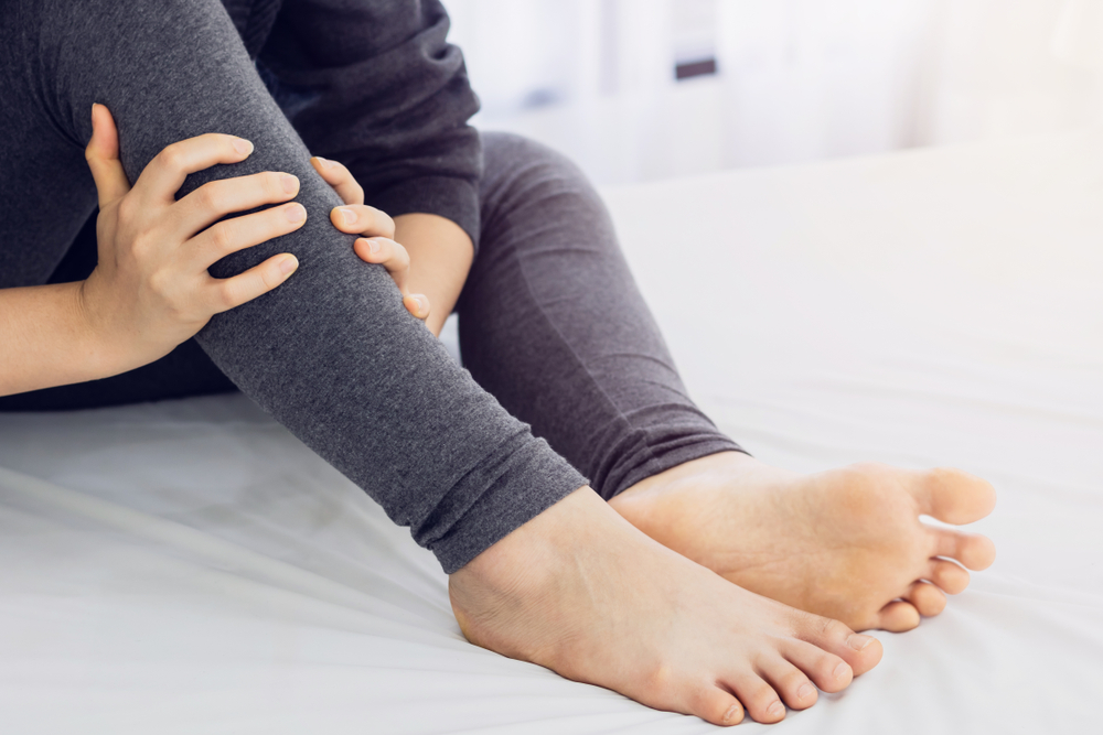 A close-up of a young adult's legs in black stretch pants. They are not wearing any shoes or socks and are clutching their right calf and shin in both hands. 