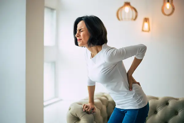 A woman in a white shirt clenches her face in pain while she grabs the lower left side of her back and leans on the side of a couch.