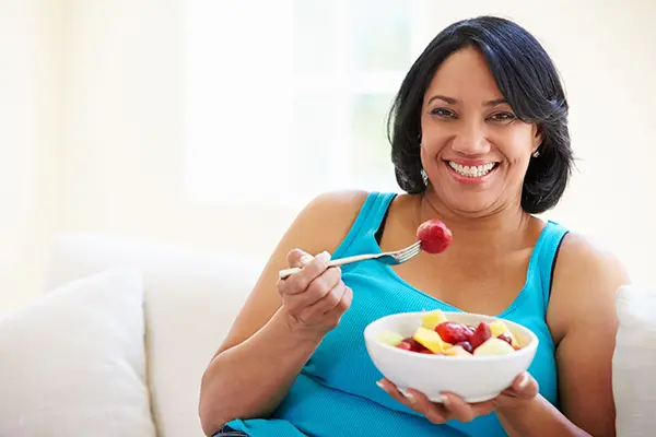 A woman in a a blue tank top and black hair, smiling at the camera as she eats a bowl of fruit