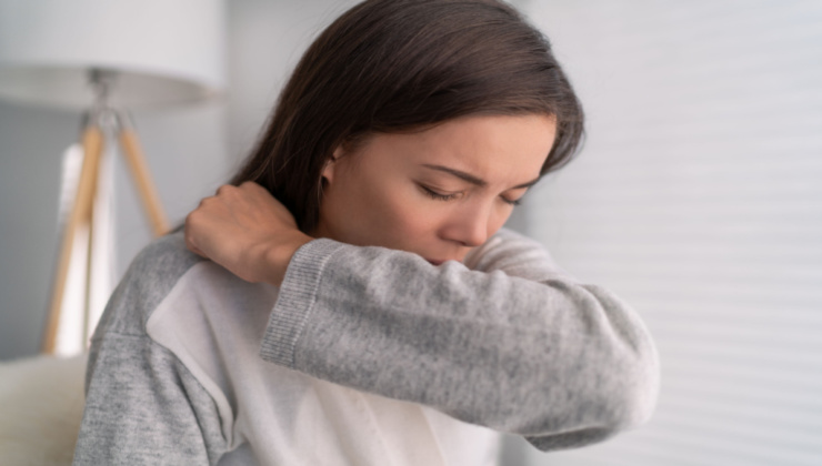 A woman with brown hair and a grey and white shirt, coughing into the crook of her elbow