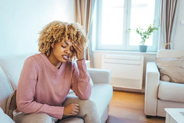 Woman sitting alone on her couch, holding her hand to her forehead and wincing, as if she is experiencing a headache. 